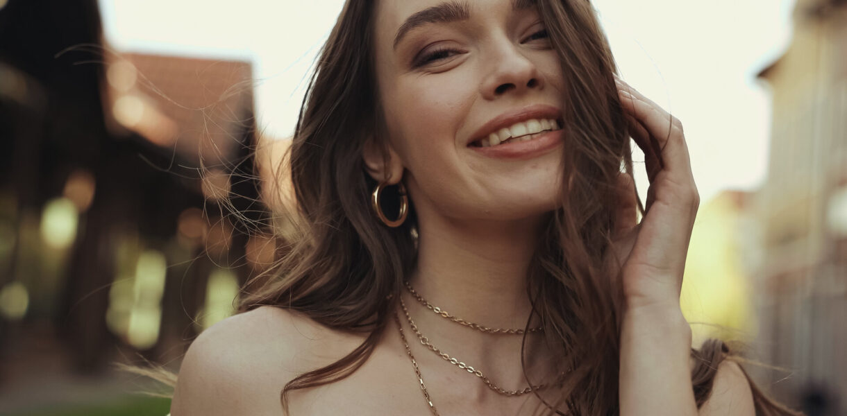 Fröhliche junge Frau mit schönem Schmuck und welligem Haar, die lächelt und in die Kamera schaut.